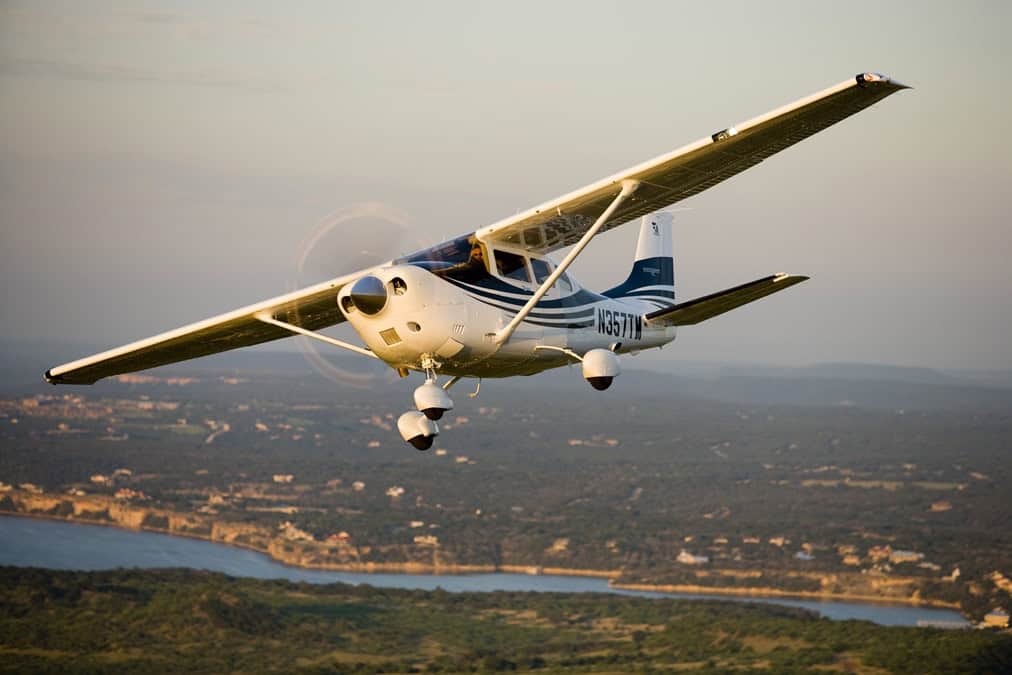 Private aircraft flying over scenic landscape with water and countryside in the background, showcasing luxury aviation services.